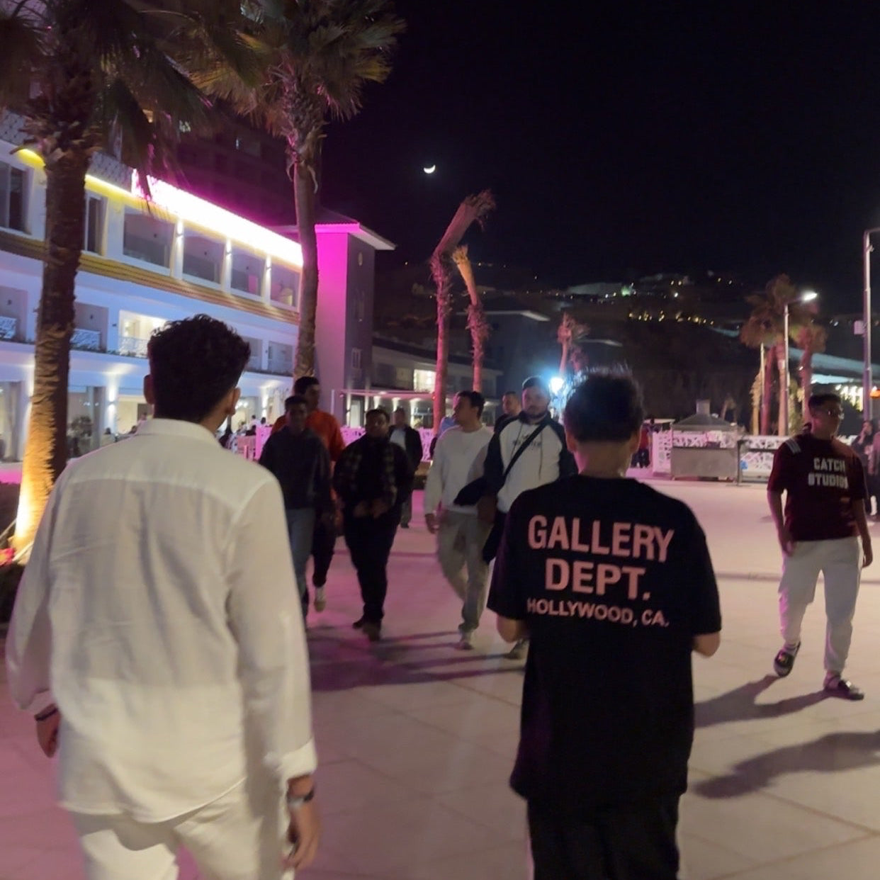 People walking on a street at night with palm trees and illuminated buildings.